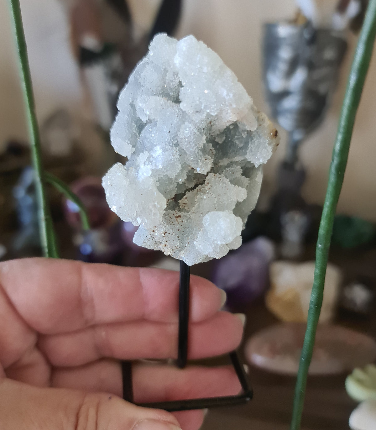 A small crystal cluster pin with a metallic appearance, held on a black pin, displayed against a backdrop of various crystal figurines and stones.