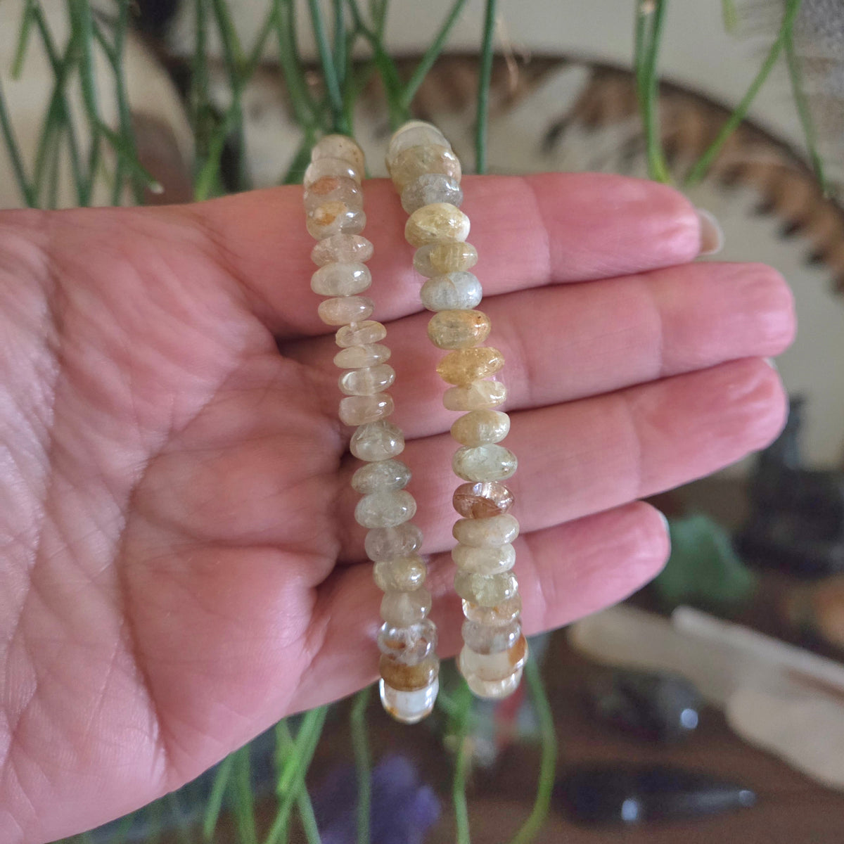 Hand holding two strands of small, round, beige beads against a blurred natural background.