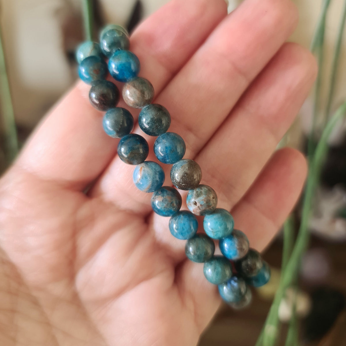 A hand holding a blue Apatite bracelet with a glossy finish, displayed against a blurred green background.