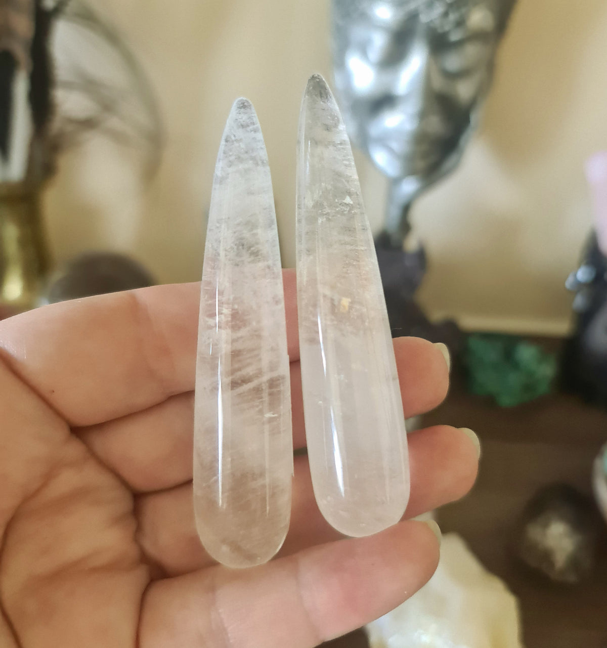Two clear quartz crystal wands being held, with a blurred background featuring various crystal figures and a statue.