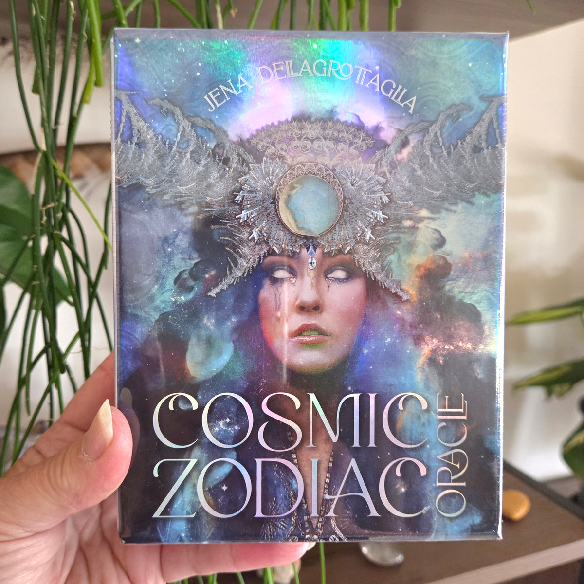 A hand holds a boxed card deck titled 'Cosmic Zodiac Oracle' by Jena Delagrotaglia. The box features an iridescent, cosmic design with a zodiac-themed illustration of a woman wearing a large ornate headdress against a galaxy background.