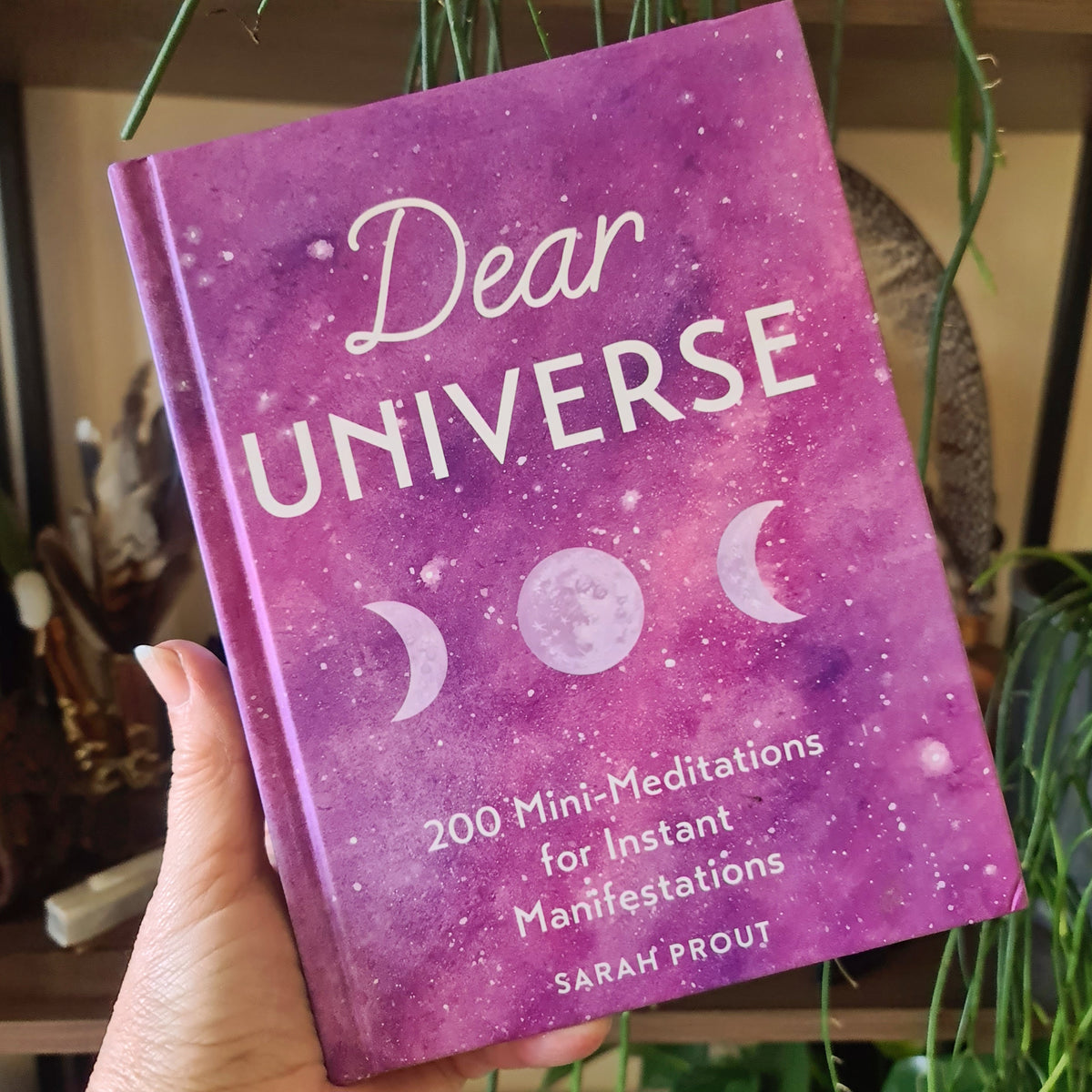 A hand holding a book titled 'DEAR UNIVERSE' by Sarah Prout, featuring a purple cover with moon and stars graphics.