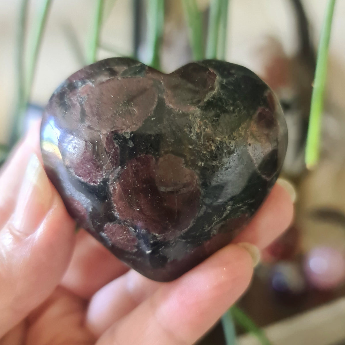 A hand holding a polished garnet crystal with a heart-like shape, with a blurred green background.