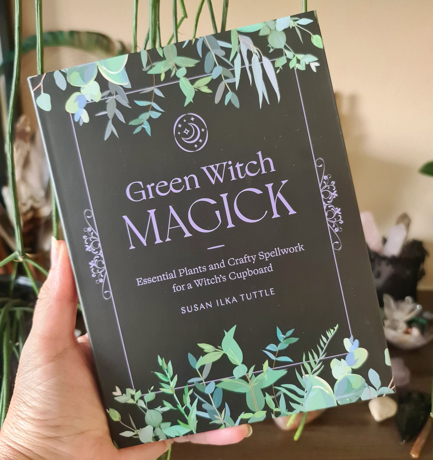 A book titled 'Green Witch Magick' by Susan Ilka Tuttle, featuring a black cover with green and purple accents and a graphic of a crescent moon and leaves.