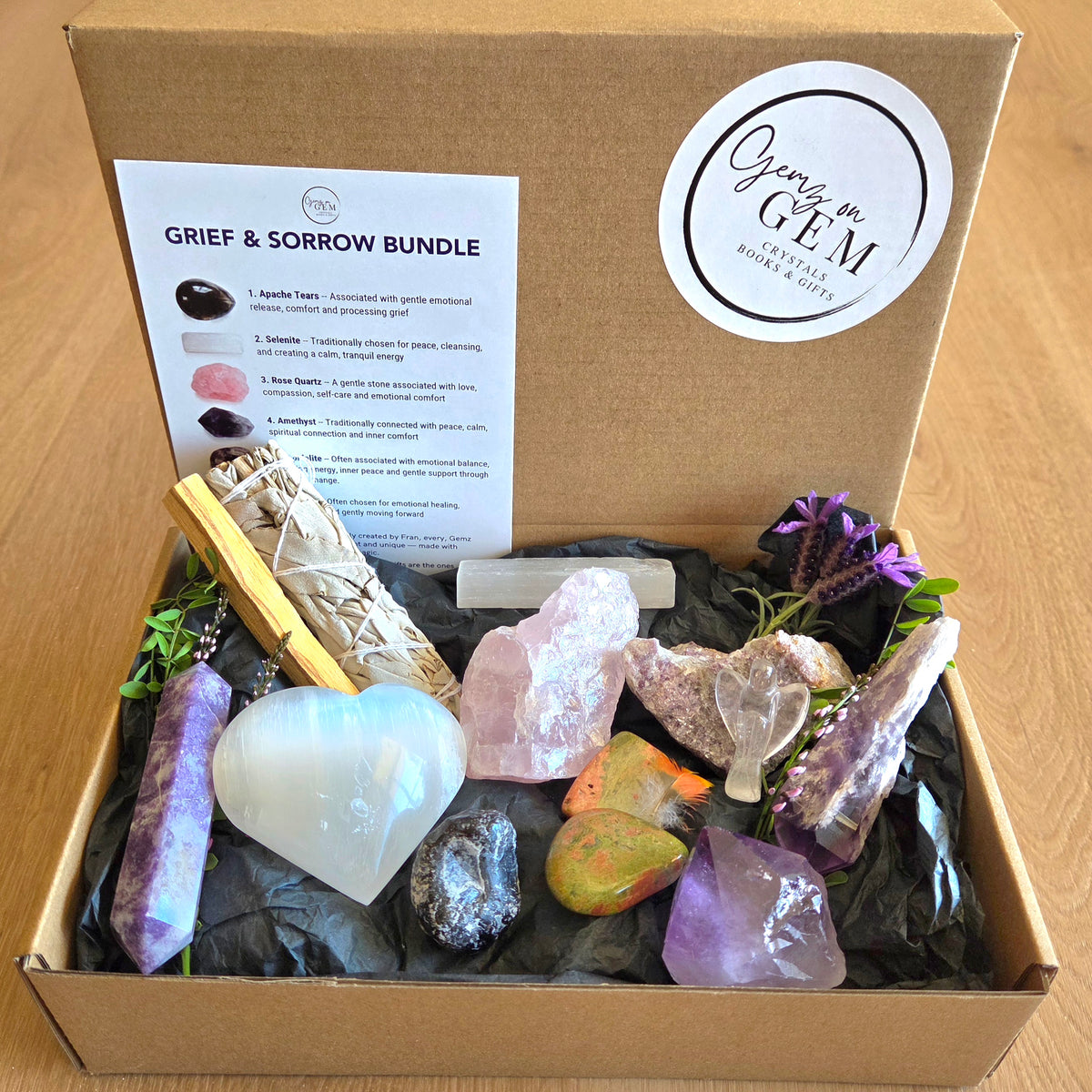An open cardboard gift box labeled 'Grief & Sorrow Bundle' by Gemz on Gem, filled with crystals, a smudge bundle, a white heart-shaped stone, a wooden wand, and a printed guide card on black tissue paper, set on a wooden surface.