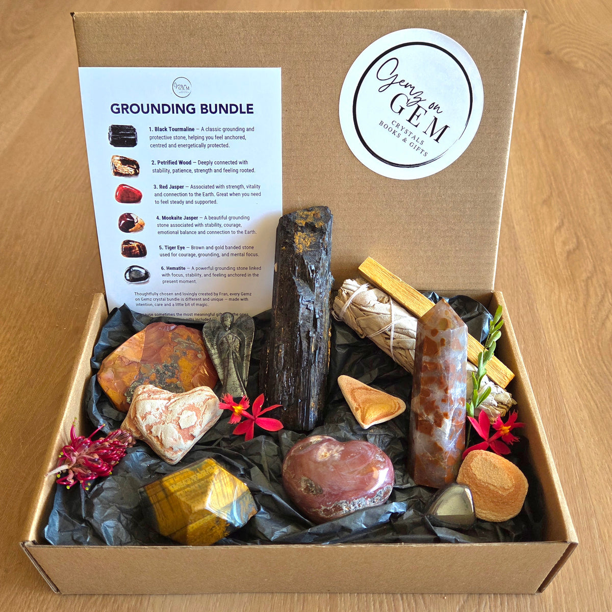 A cardboard gift box filled with a grounding crystal bundle, including polished stones, a black wood cylinder, a wrapped bundle, and a printed guide card titled 'Grounding Bundle.' The box lid has a round logo reading 'Gem & Gem.