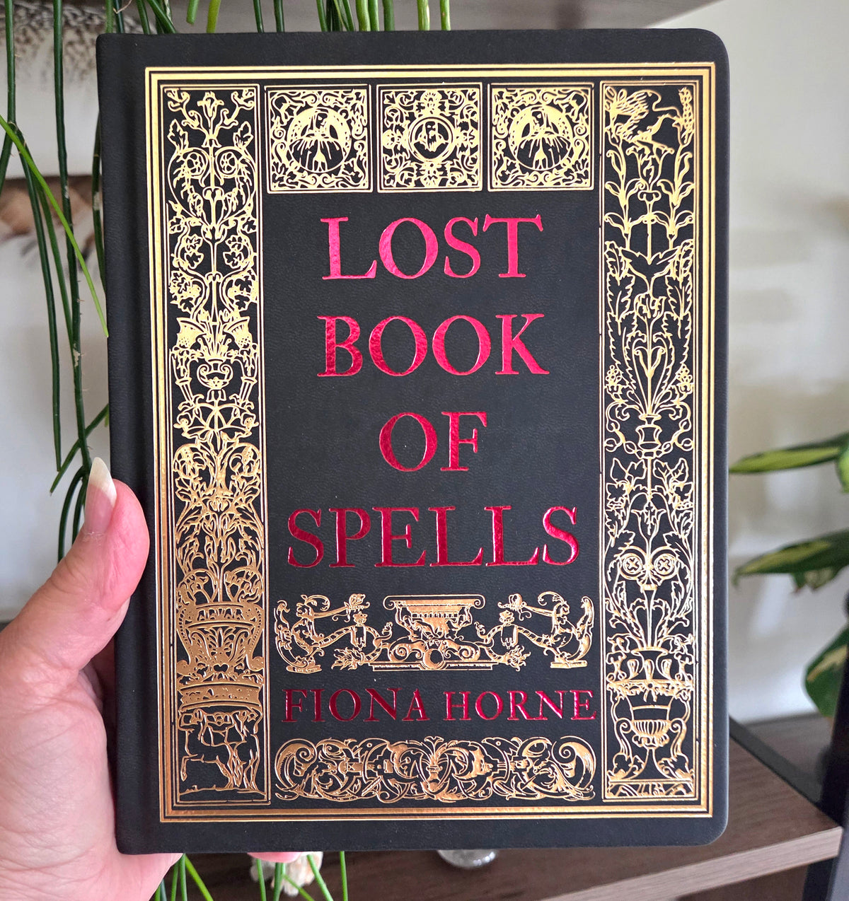 A hand holds a hardcover book titled 'Lost Book of Spells' by Fiona Horne. The book cover is black with ornate gold filigree borders and the title and author name in red text.