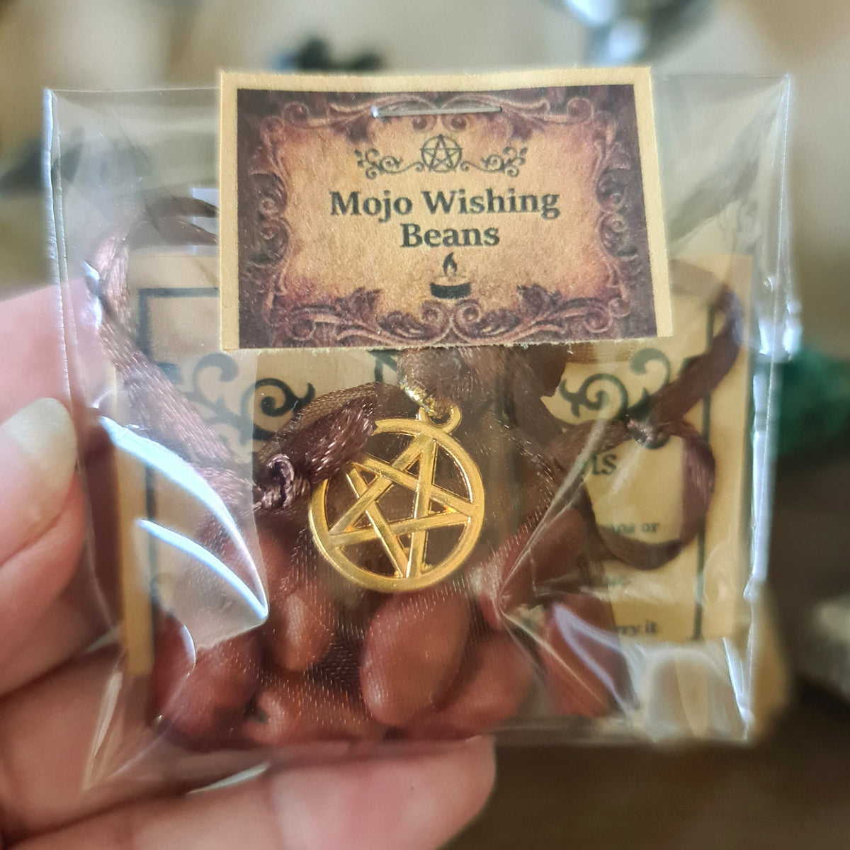 A pouch containing seven large, brown mojo wishing beans with a gold pentagram charm, packaged in a clear plastic bag with a label reading 'Mojo Wishing Beans'.