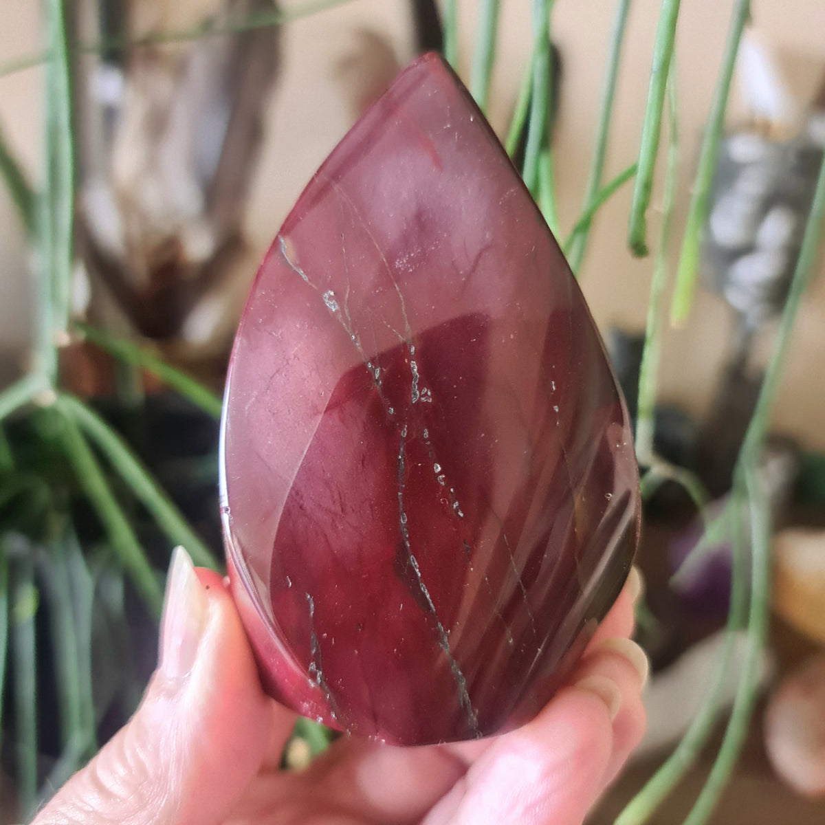 A polished red Mookaite Jasper Flame held in a person's hand, with a blurred background suggesting indoor setting