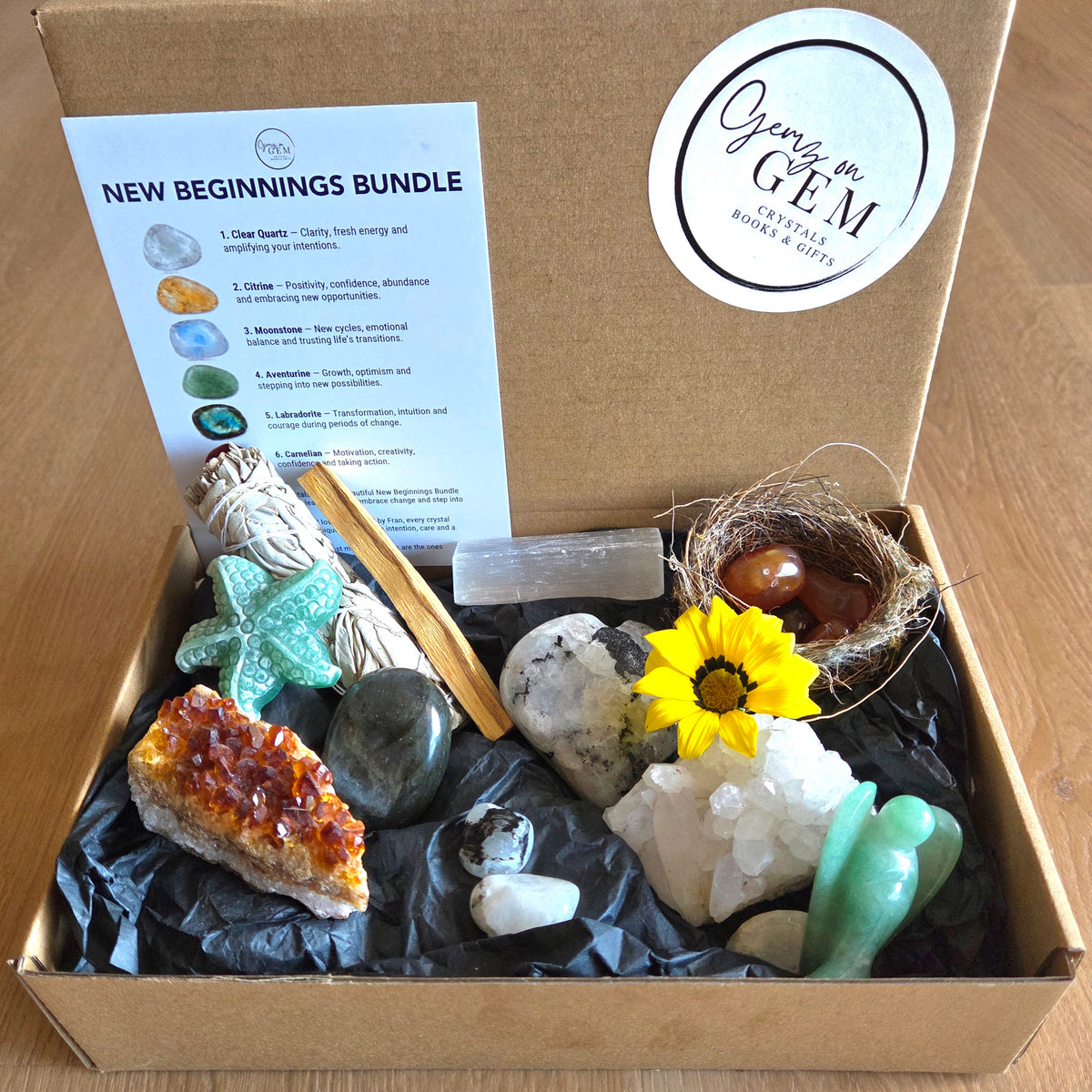 A Geng GEM 'New Beginnings Bundle' gift box filled with various crystals, stones, a small bird nest with two brown eggs, a yellow sunflower, and a printed guide card explaining the bundle contents.