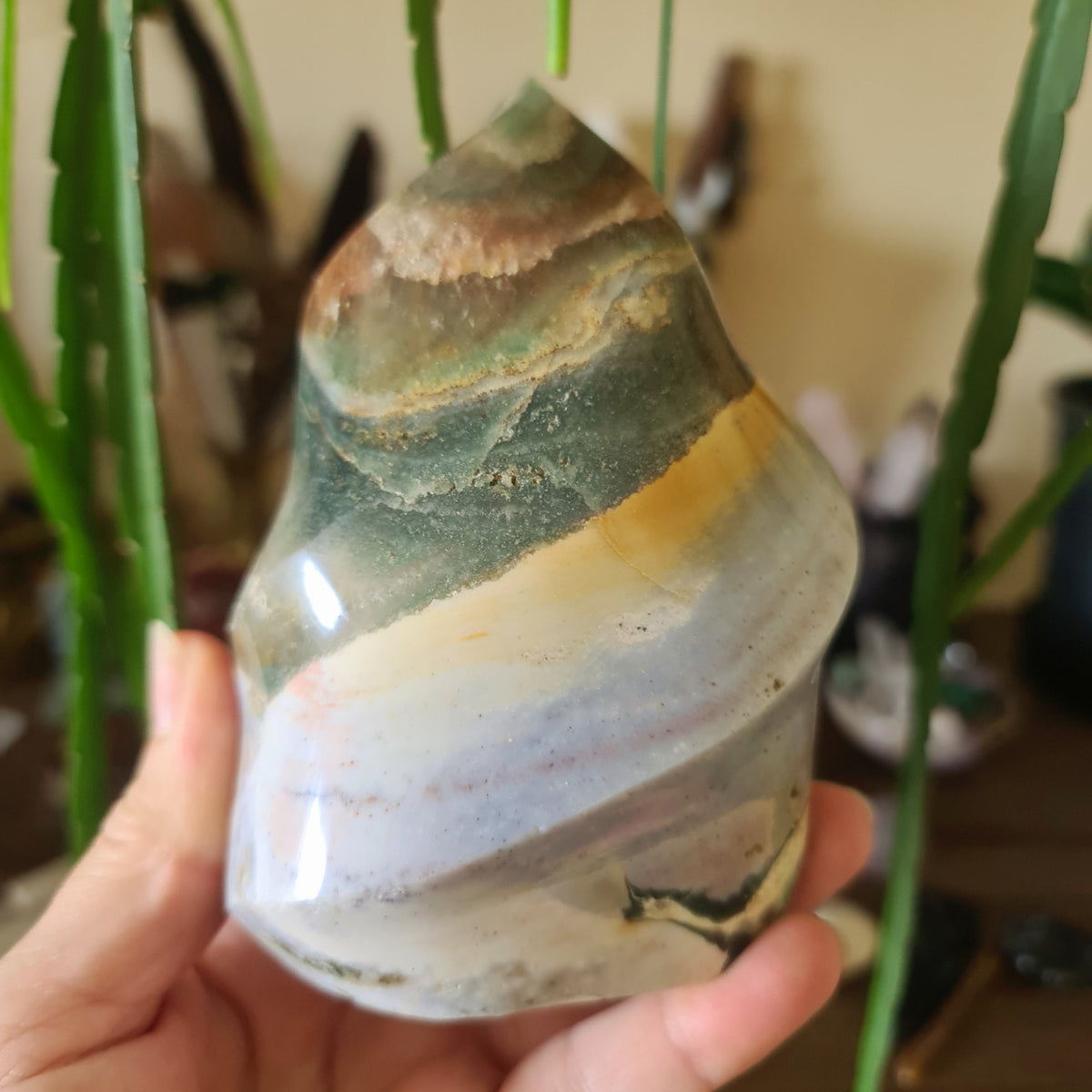 A hand holding a polished Ocean Jasper crystal with a flame-like shape, with colors ranging from green, brown, and white.
