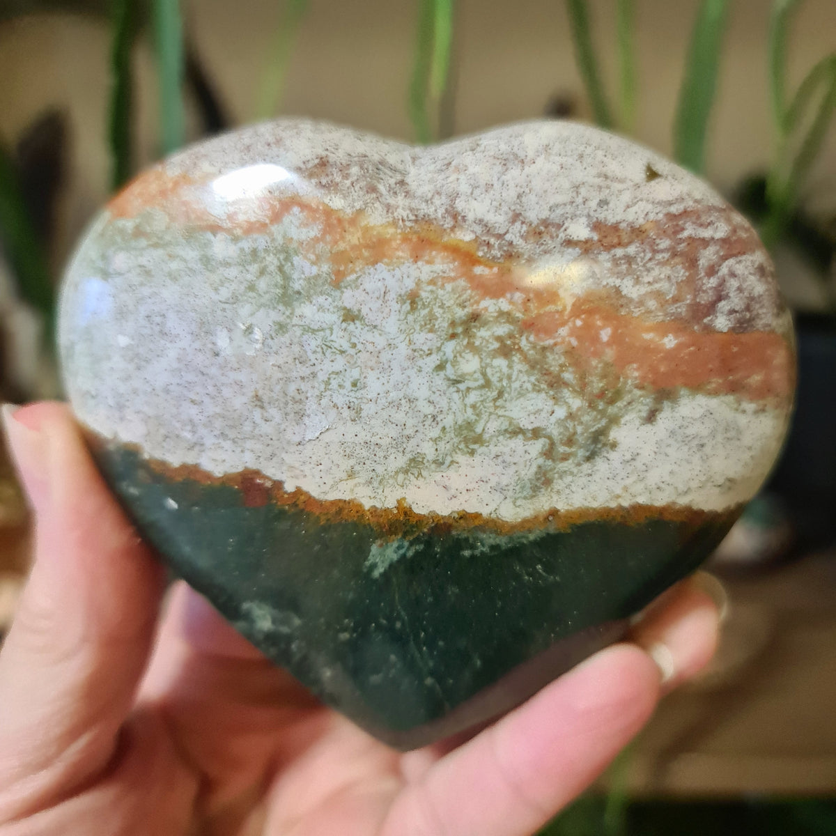 A hand holding a large heart-shaped Ocean Jasper stone with green, brown, and white color patterns.