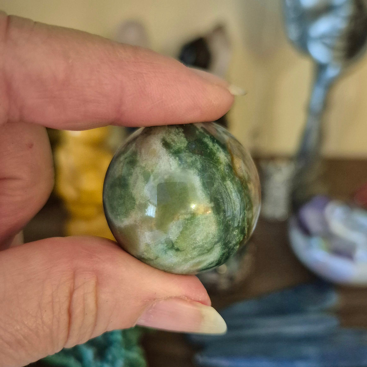 A small Ocean Jasper sphere held in a person's hand, with a blurred background containing various colorful items.