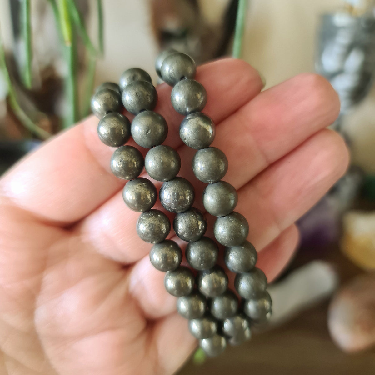 A person's hand holding a series of round pyrite crystal beads, with a plant and various items in the background.