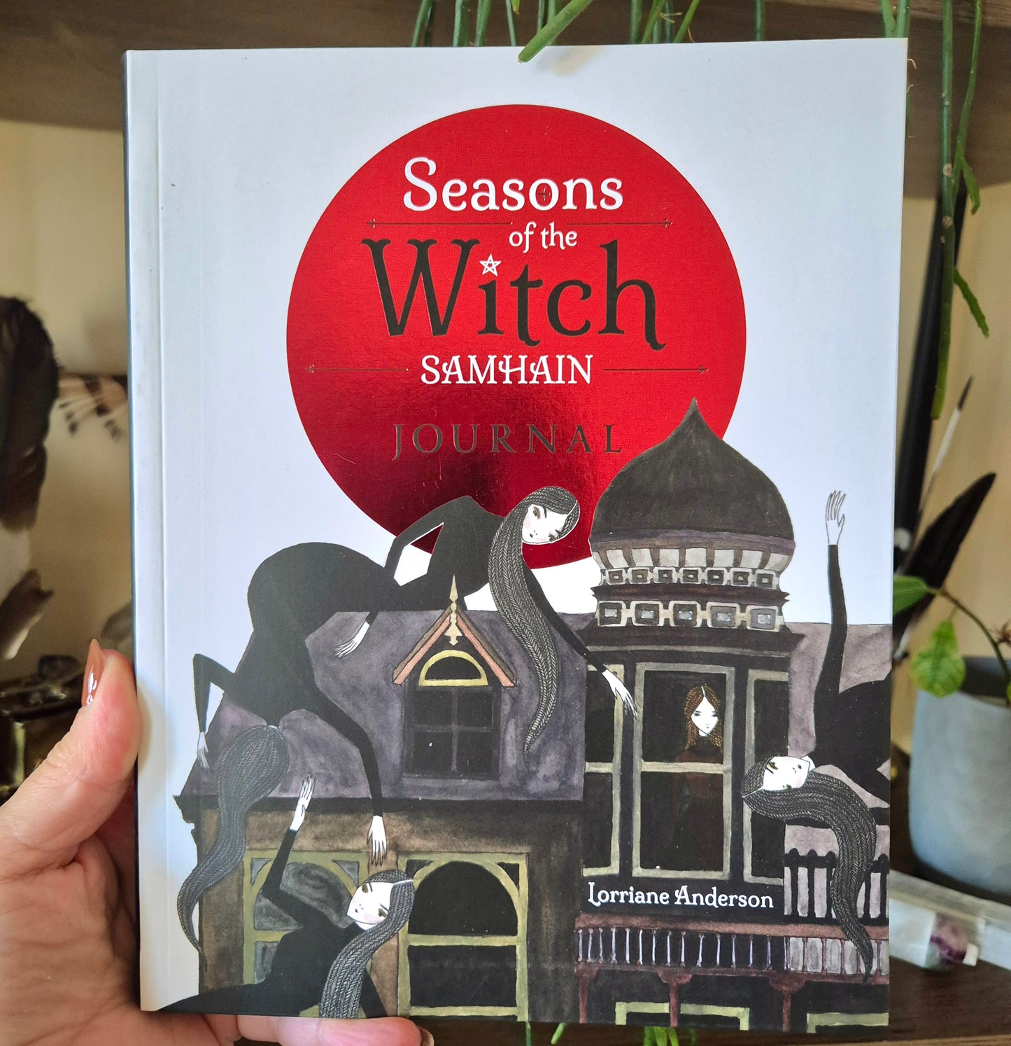 A hand holding a book titled 'Seasons of the Witch Samhain Journal' with a red and black cover design featuring a witch's house and a full moon.