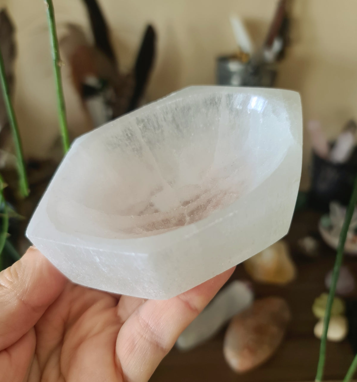 A clear, flat, bowl-shaped selenite crystal holder, held in a person's hand, with a blurred background suggesting a home or personal item.