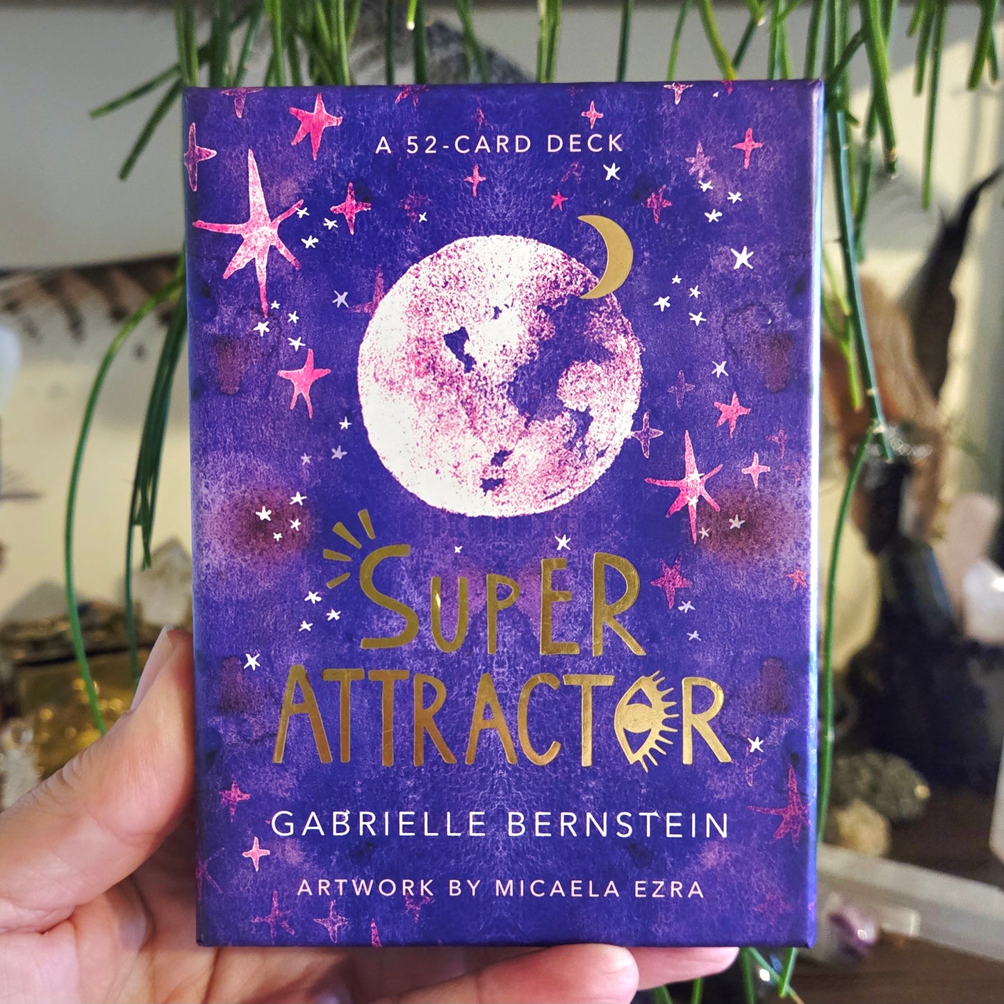 A person holding a pack of 'Super Attractor' oracle cards by Gabrielle Bernstein with artwork by Micaela Ezra. The packaging is predominantly purple with images of a moon and stars.
