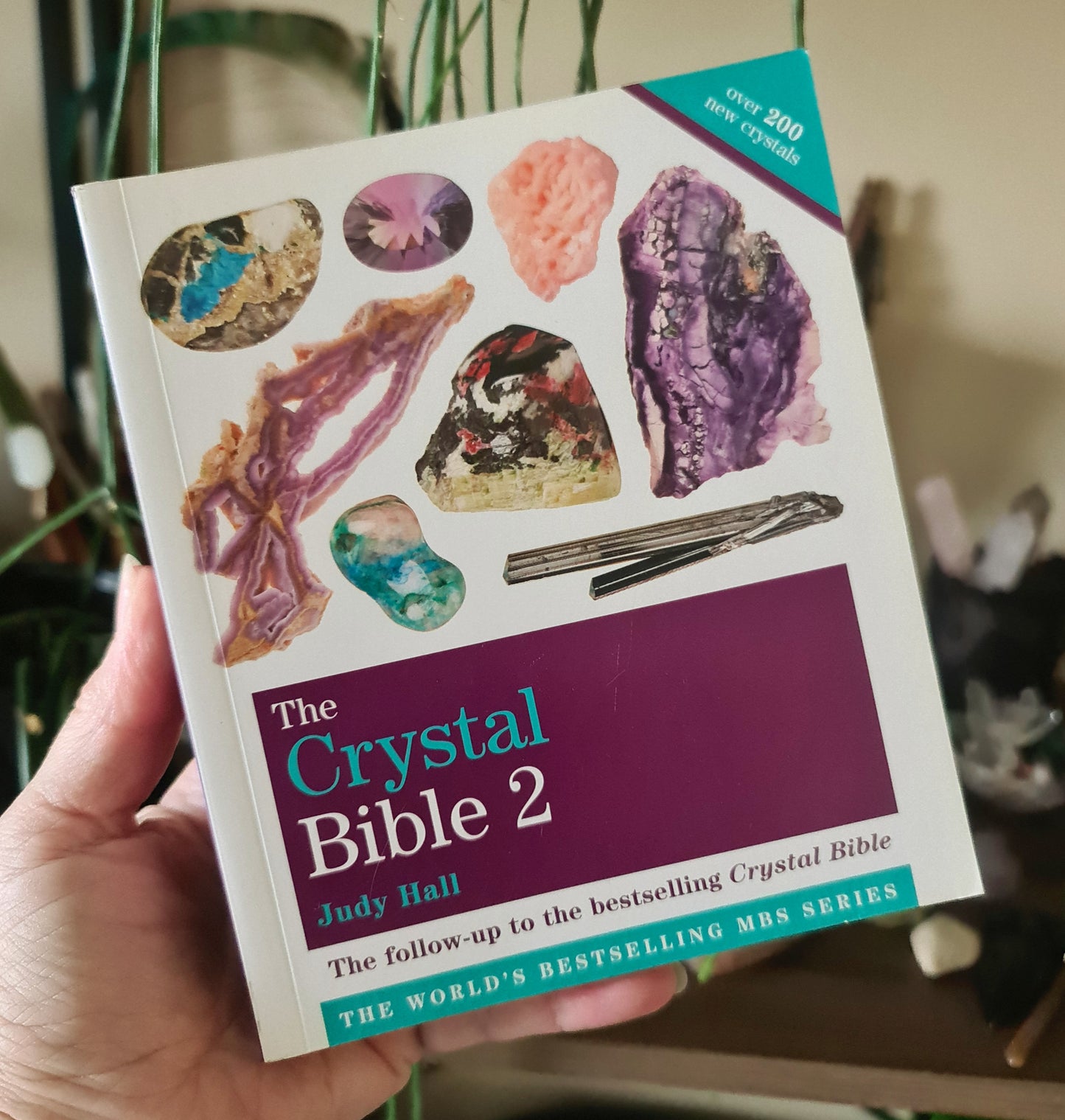 A hand holding a purple book titled 'The Crystal Bible 2' by Judy Hall, which is a part of the Crystal Ms Series.
