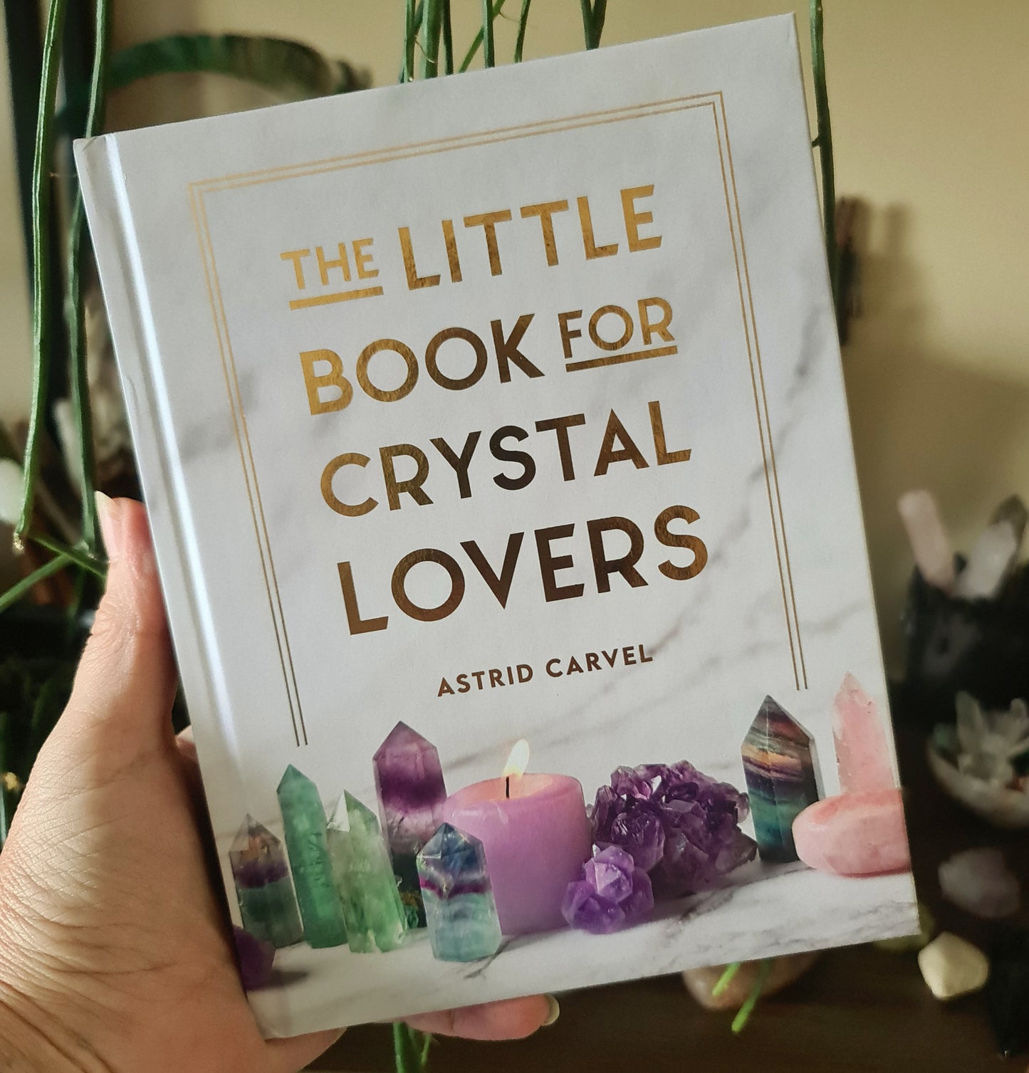 A hand holding a book titled 'The Little Book For Crystal Lovers' by Astrid Carvel, featuring a marble-like background and an image of a candle with crystals on the cover.