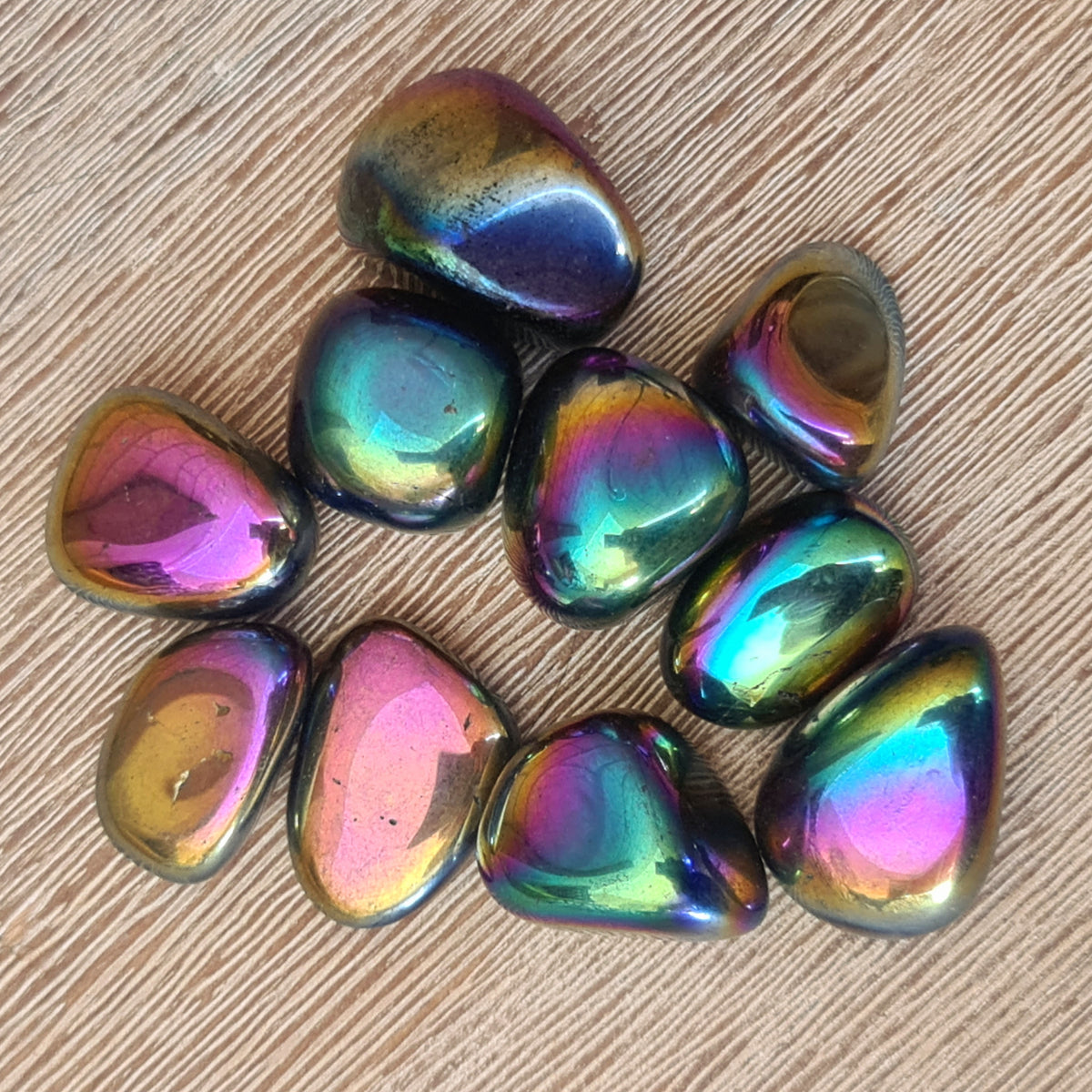 A collection of multicolored titanium aura tumble stones with a metallic sheen, displayed on a wooden surface.