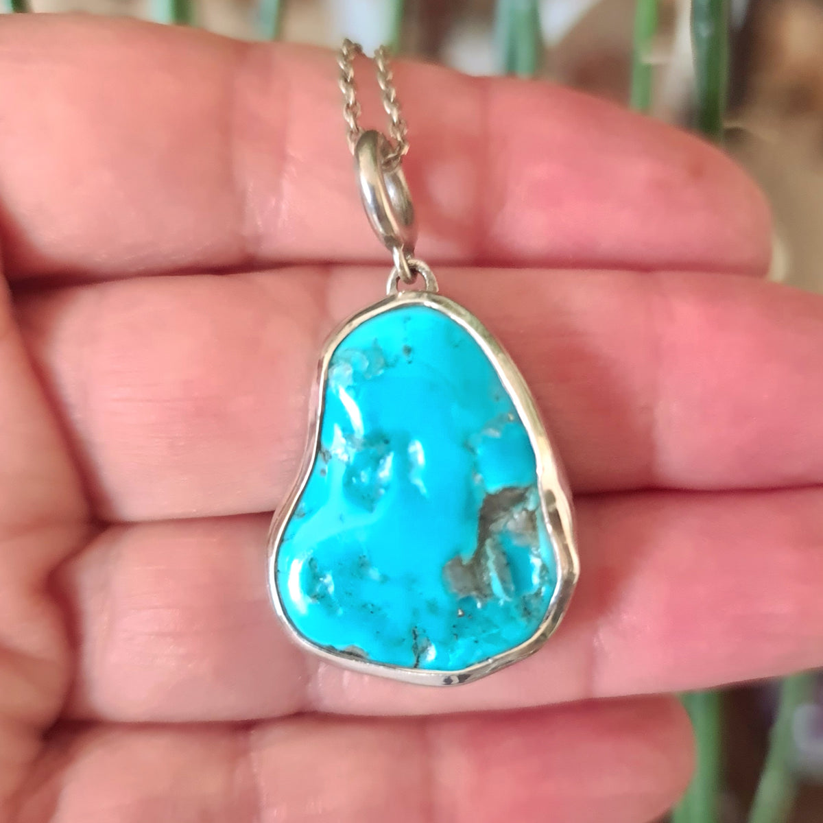 A sterling silver pendant with a turquoise stone held in a hand, with a plant in the background.