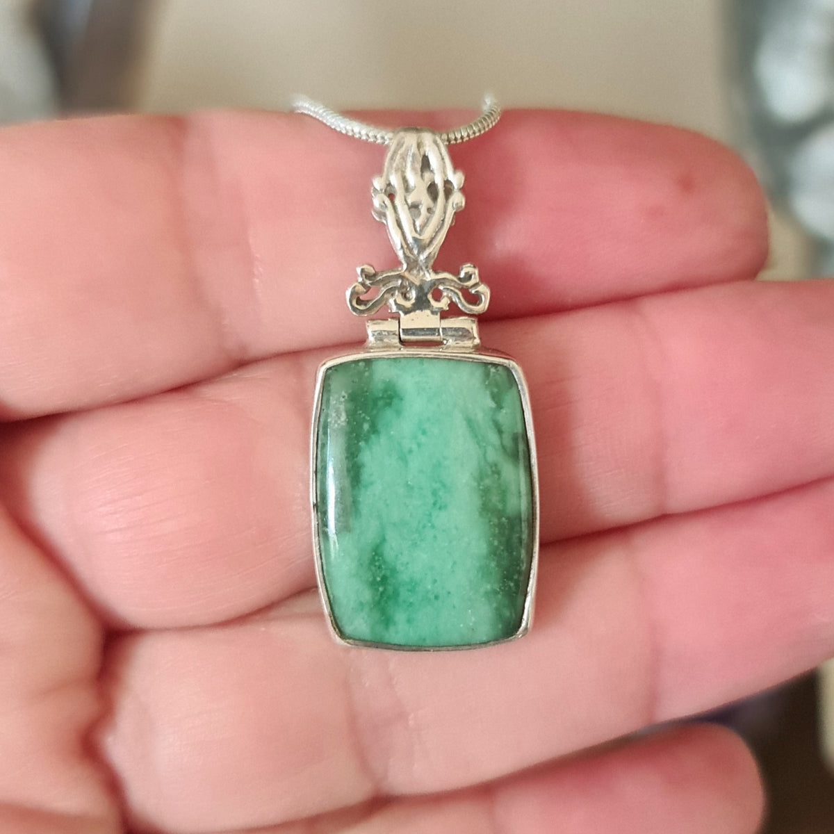 A hand holding a sterling silver pendant with a green variscite gemstone set in it, featuring intricate metalwork on the top border.