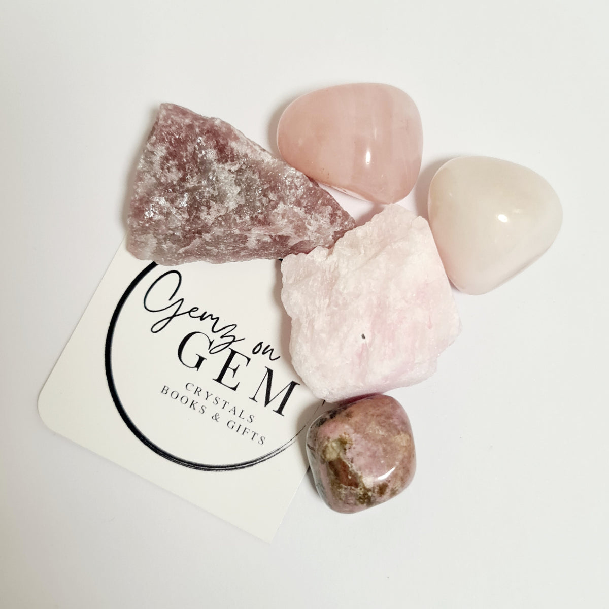 An assortment of pink and rose-colored crystals, including tumbled stones and raw crystal pieces, displayed on a branded card with 'Gemz on Gem' printed on it.