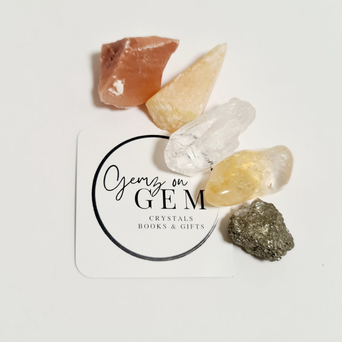 An assortment of polished crystals including Honey Calcite, Citrine, Red Calcite, Pyrite, and Clear Quartz, displayed on a white background with a business card that reads 'Gemz on Gem'.