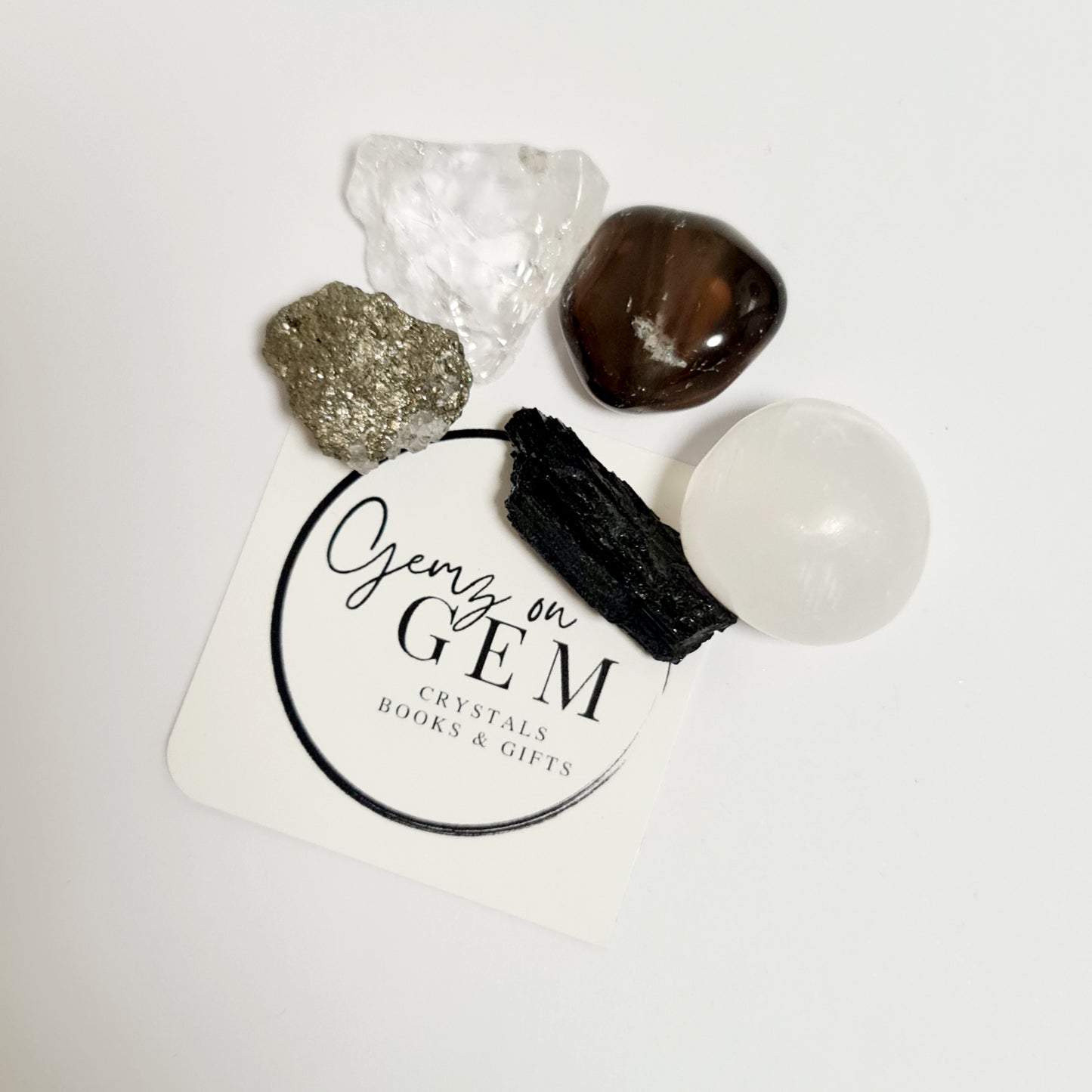 An assortment of polished and unpolished gemstones on a branded card, including a selenite, clear quartz, pyrite, smoky quartz, and black tourmaline.