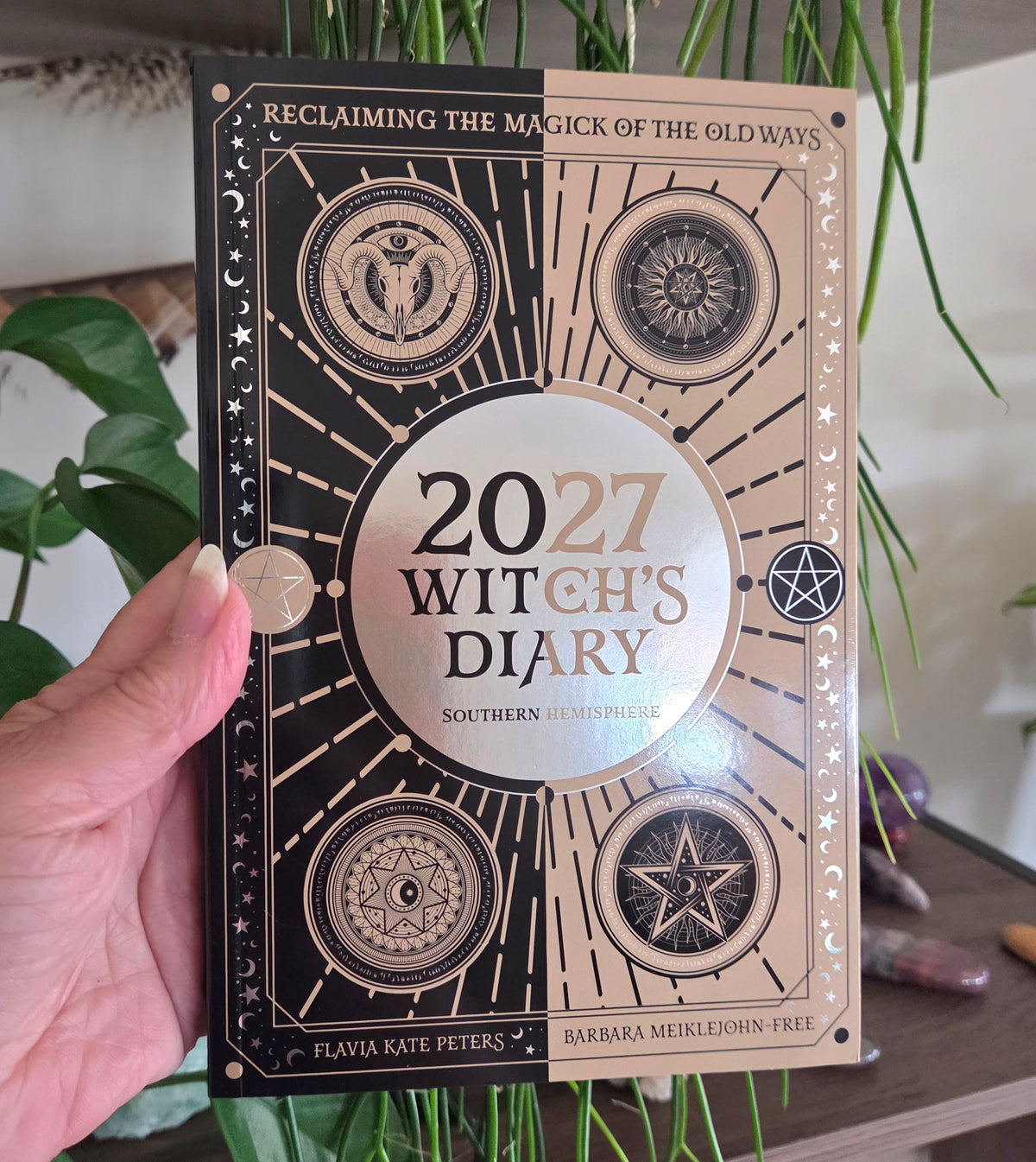 A hand holds a hardcover journal titled '2027 Witch's Diary' against a background of green houseplants and crystals. The cover features a split black and beige design with gold celestial illustrations.