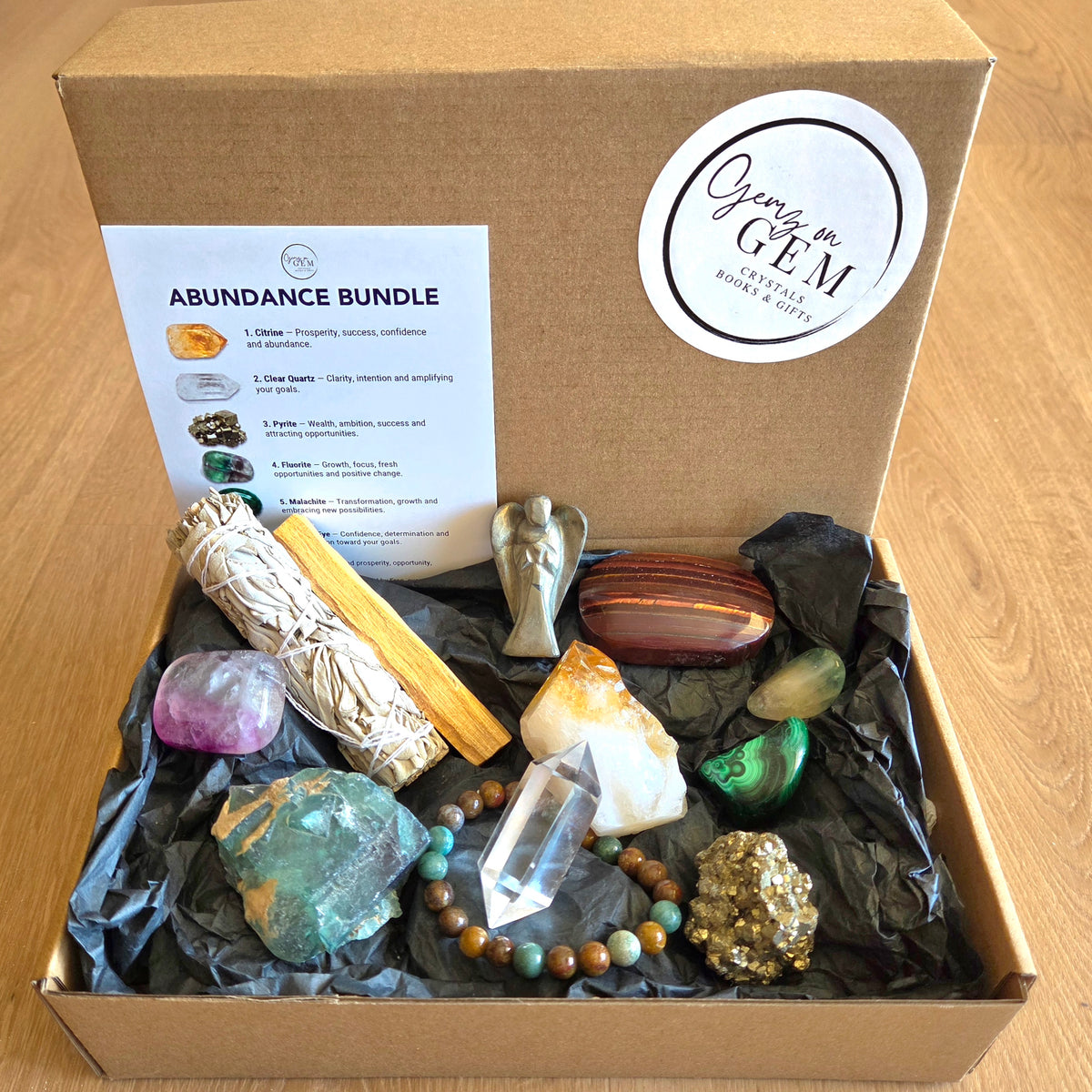 An 'Abundance Bundle' gift box from Gently in GEM, containing a smudge bundle, rose quartz, clear crystal, green aventurine, brown and white stones, a silver angel, a striped red and brown stone, a green malachite piece, and a beaded bracelet, all arranged in black tissue paper inside an open kraft cardboard box.