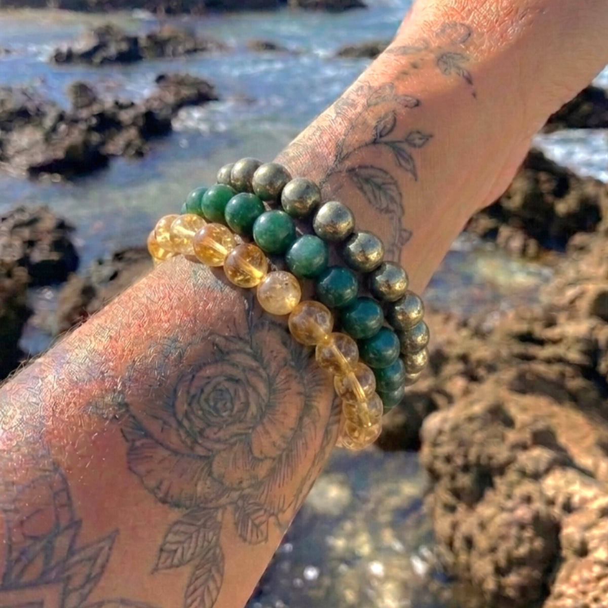 Three beaded bracelets worn on a tattooed wrist, featuring green stone beads, metallic gold beads, and translucent amber-yellow beads, set against a rocky beach background.