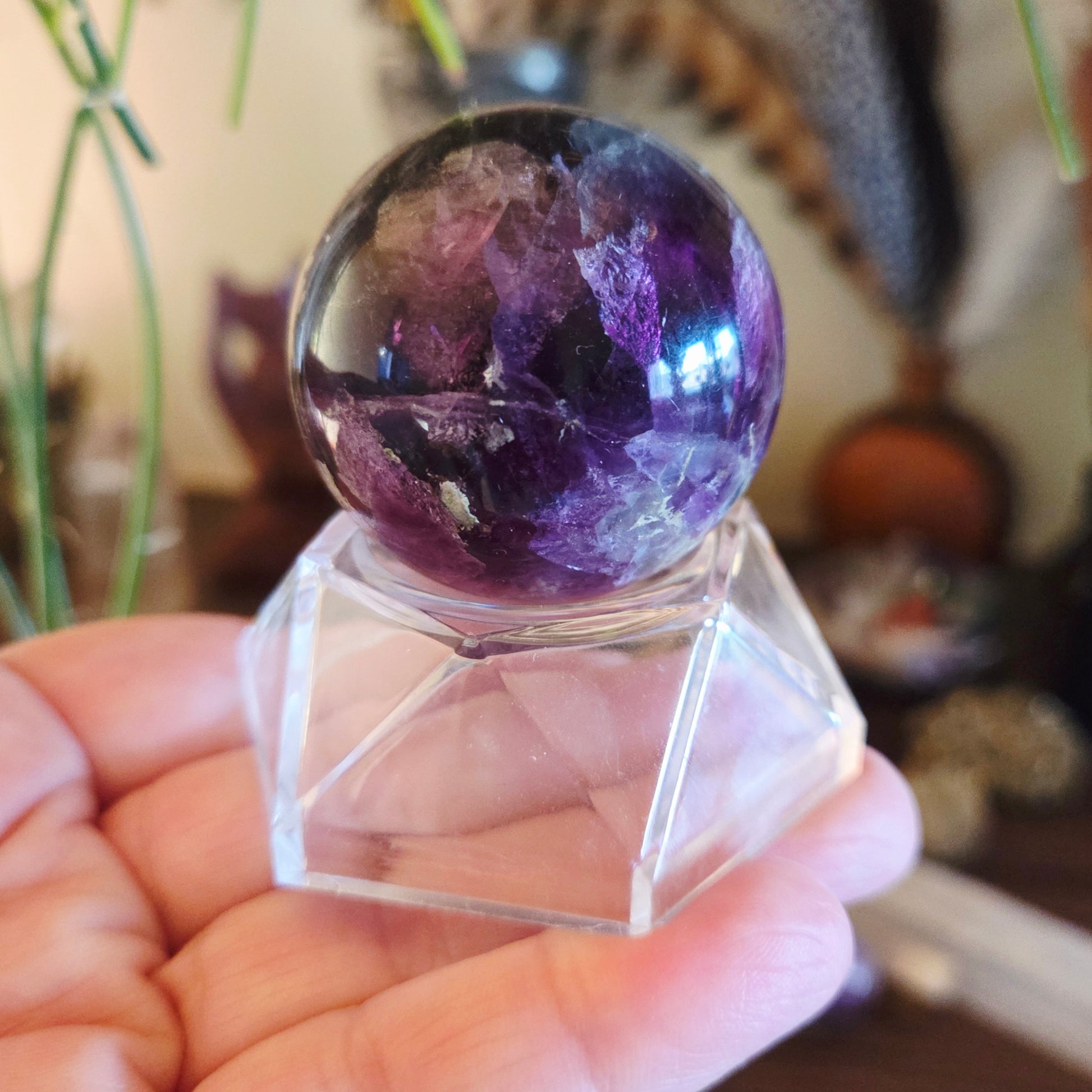 A hand holding a purple sphere-shaped crystal on a clear acrylic stand.