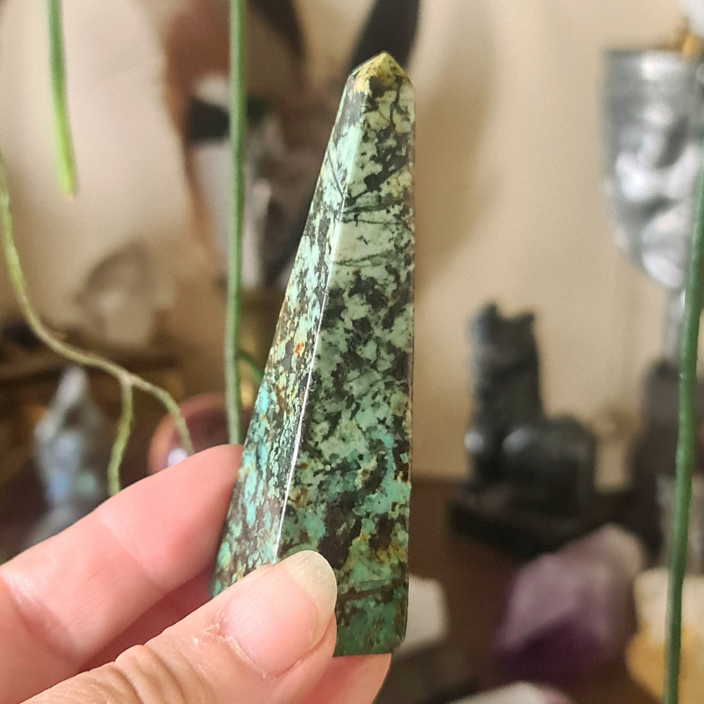 A hand holding a small turquoise obelisk-shaped crystal figure, with a blurred background featuring other crystal figures and plants.