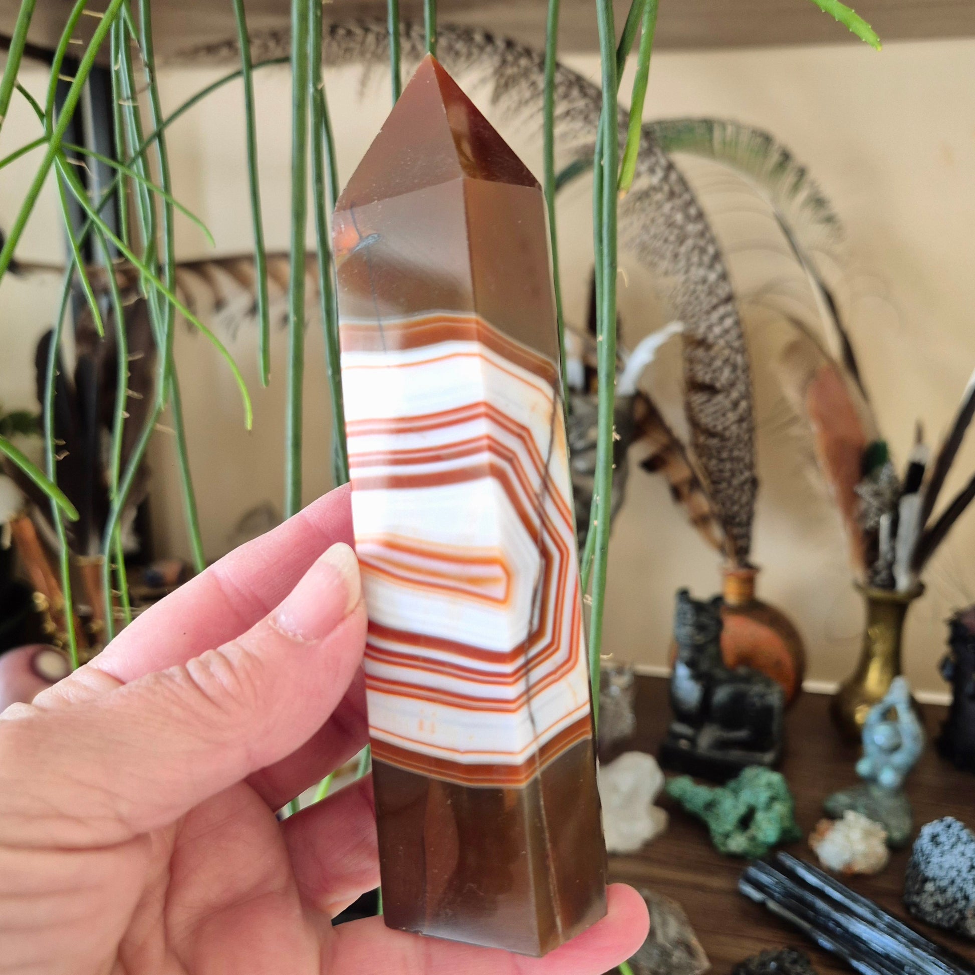 A hand holding a agate tower with a striped pattern, with a plant in the background and various other crystal figurines.