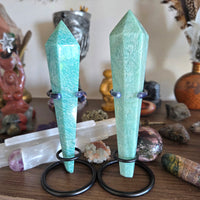 Two green crystal points on stands with a blurred background of crystals and statues.