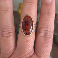 A silver band ring featuring an oval cabochon gemstone with a rich amber-brown color and fiery orange inclusions, shown worn on a person's finger.