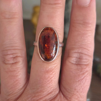 A silver band ring featuring an oval cabochon gemstone with a rich amber-brown color and fiery orange inclusions, shown worn on a person's finger.