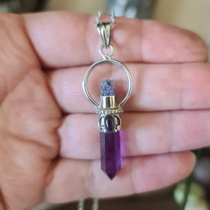 A hand holding a purple amethyst, aqua aura, and garnet crystal pendant with a silver cap and ring on top.