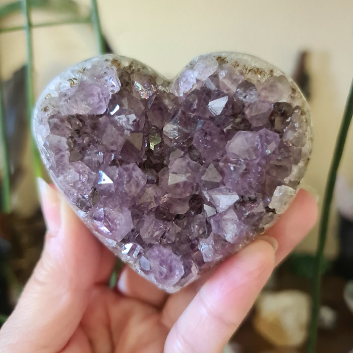 A hand holding a heart-shaped amethyst crystal cluster with multiple facets and a purple coloration.