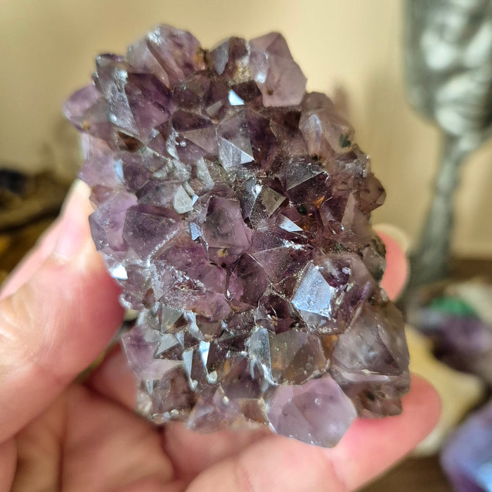 A large Amethyst crystal cluster held in a person's hand, with a blurred background.