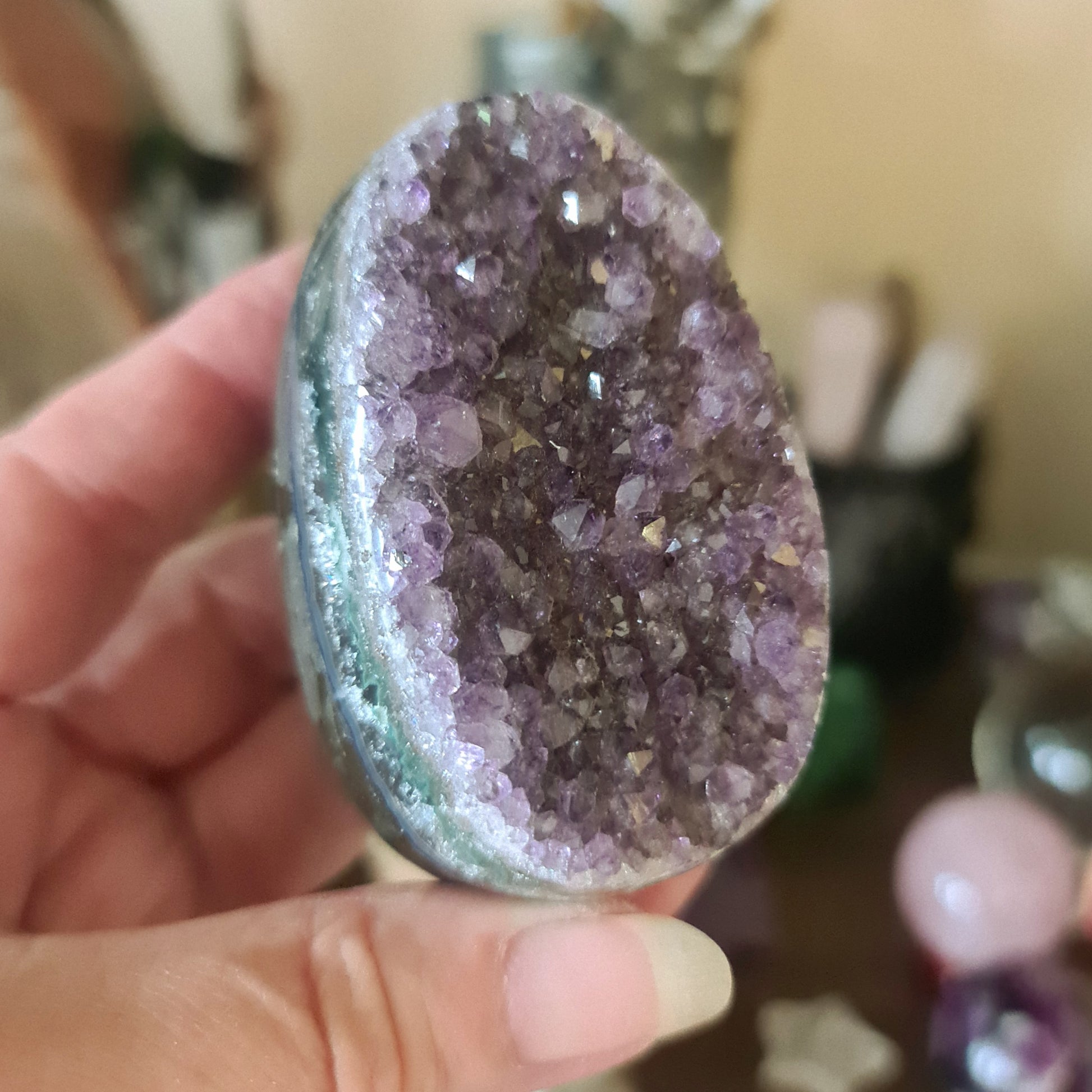 A polished amethyst geode egg with a cracked surface, held in a hand, against a blurred background of similar decorative items.