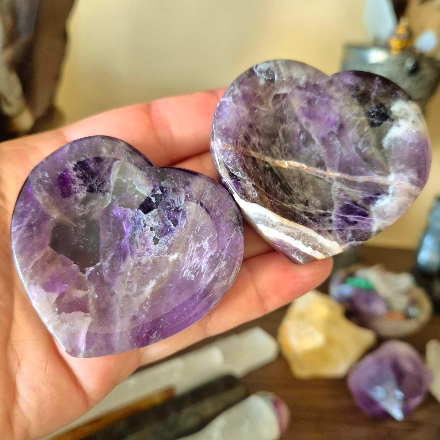 Two heart-shaped amethyst trinket bowls held in a person's hand, with a blurred background featuring other gemstones and decorative items.