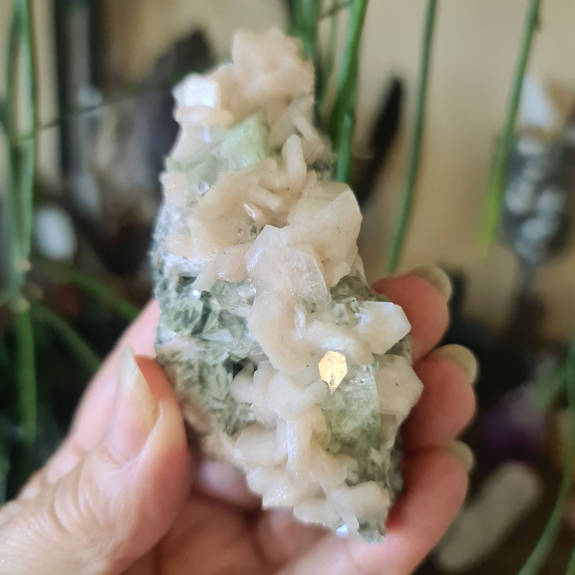 A small crystal cluster on a stand, predominantly green with white and pink tones, held in a person's hand.