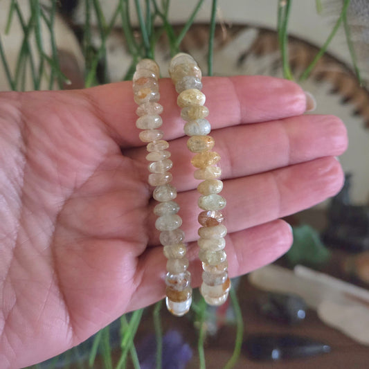 Hand holding two strands of small, round, beige beads against a blurred natural background.