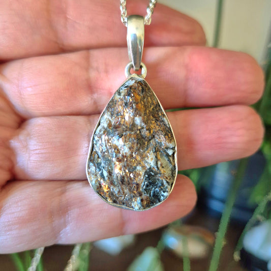 A hand holding a silver pendant with a metallic and reflective surface, set against a blurred background.