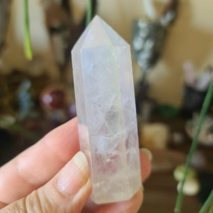 A hand holding a polished, clear Aura Quartz crystal point with a tapered shape, against a backdrop of various items.