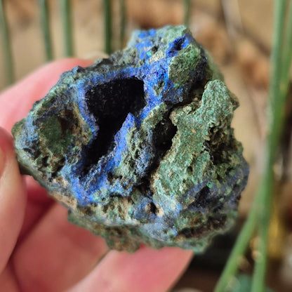 A raw Azurite Malachite mineral specimen held in a person's hand, displaying its natural rugged texture and typical green color with hints of blue.