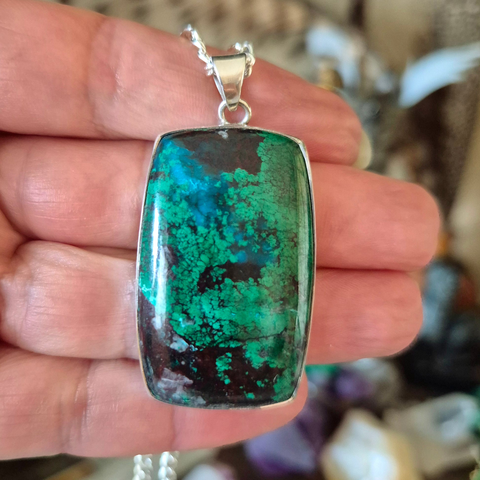 A blue and green azurite malachite pendant held in a hand, attached to a silver-colored chain.