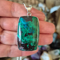 A blue and green azurite malachite pendant held in a hand, attached to a silver-colored chain.